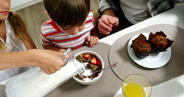 Children are pouring milk into a bowl of breakfast cereal with strawberries at the family breakfast table. A plate with muffins and a glass of orange juice is nearby. This image is ideal for promoting healthy eating habits, family bonding moments, morning routines, and breakfast foods.