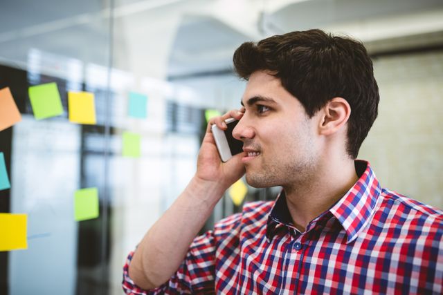 Smiling businessman talking on phone while looking at sticky notes attached in creative office