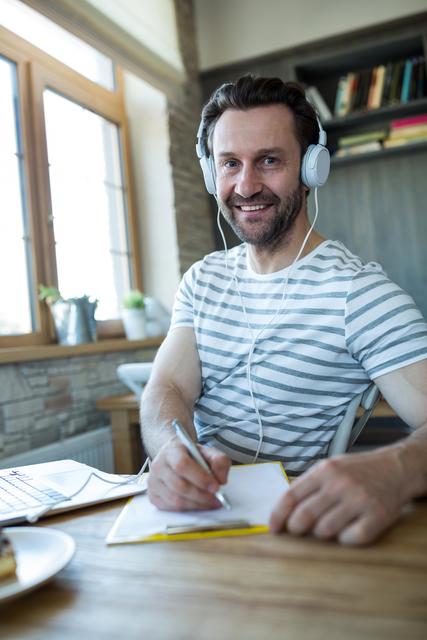 This image depicts a man sitting at a table in a coffee shop, wearing headphones and writing in a diary. He is smiling and appears relaxed, suggesting a casual and enjoyable moment. The natural light from the window and the cozy interior create a warm atmosphere. This image can be used for promoting lifestyle blogs, articles about journaling, creative writing, or advertisements for coffee shops and headphones.