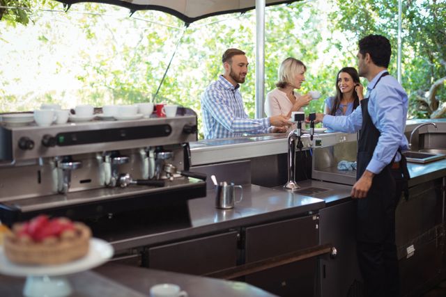 Waiter serving coffee to customers at an outdoor cafe counter. The scene depicts a friendly interaction between the waiter and the customers, highlighting excellent customer service. Ideal for use in articles or advertisements related to hospitality, customer service, cafes, restaurants, and social interactions.