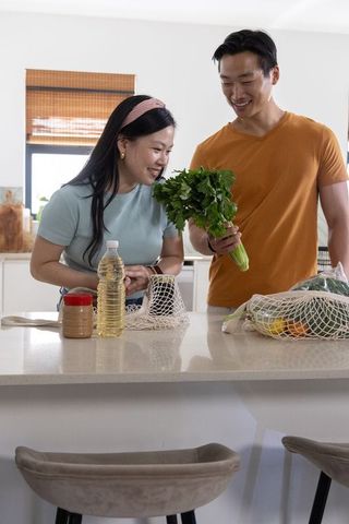 Asian couple unpacking groceries at modern kitchen island, smiling while checking herbs