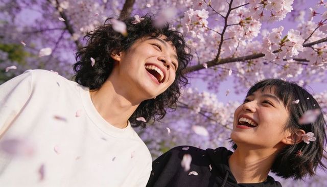 Young couple laughing under cherry blossoms in spring park, petals falling, joyful moment