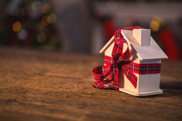 Toy house wrapped with red ribbon on wooden table, creating a festive and cozy atmosphere. Ideal for holiday-themed promotions, Christmas cards, home decor inspiration, and gift ideas. Perfect for conveying warmth and celebration during the holiday season.