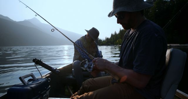 Two men are fishing in a small boat on a calm lake surrounded by mountains during sunrise. They are wearing hats and casual clothes, focusing on their fishing rods. Ideal for use in content related to fishing, outdoor recreation, summer activities, travel adventures, and peaceful nature scenes.