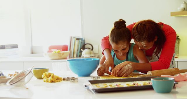 Mother and daughter baking cookies in home kitchen with flour-covered hands. Daughter concentrates on shaping dough while mother assists from behind. Ideal for illustrating family bonding, cooking activities, parent-child relationships, and home lifestyle themes.