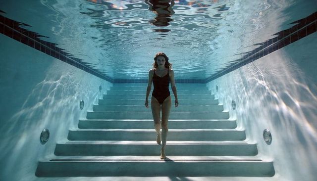Woman descending underwater pool stairs with serene symmetry and light caustics