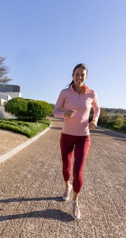 Female jogging on sunny park path in pink top and maroon leggings for healthy running