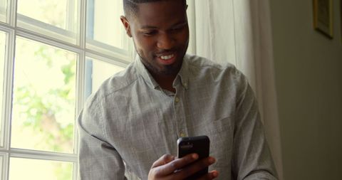 Young Man Smiling While Using Smartphone by Window