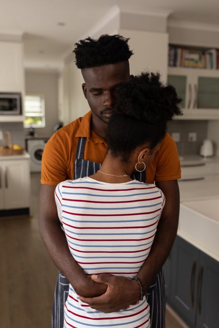 Loving african american young man wearing apron with eyes closed embracing girlfriend in kitchen. romance, unaltered, lifestyle, home, love and togetherness concept.