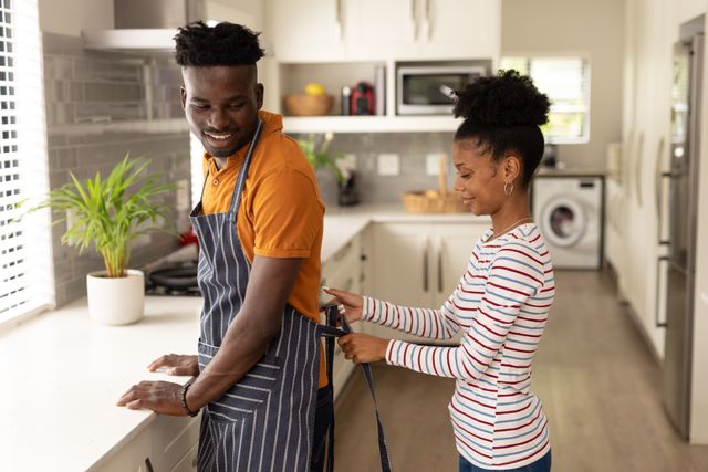Young African American couple enjoying time together in modern kitchen. Woman tying apron on boyfriend while they prepare a meal. Perfect for themes related to love, relationships, domestic life, and cooking. Ideal for use in lifestyle blogs, home and kitchen advertisements, and relationship advice articles.