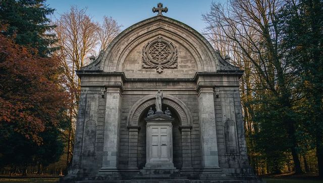 Historic stone mausoleum chapel with cross and statue amid autumn trees