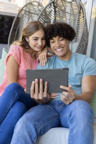 Young couple watching tablet together on sunlit porch lounge, smiling cozy casual moment