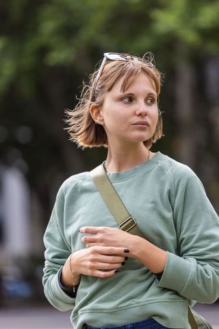 Young woman pausing in urban park wearing sage sweatshirt adjusting olive crossbody strap
