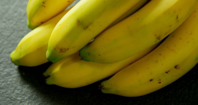 Close-up of ripe yellow bananas resting on dark tabletop. Perfect for use in advertising healthy eating, tropical produce promotions, organic food products, grocery store advertisements, and health-conscious meal planning visuals.