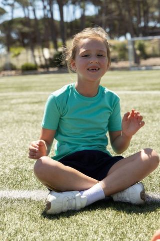 Young girl sitting cross-legged on turf smiling in teal shirt at outdoor sports practice
