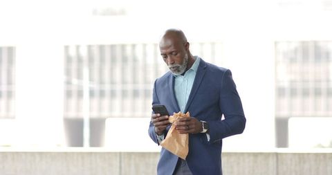 African American Executive Checking Smartphone While Eating Sandwich on Urban Rooftop
