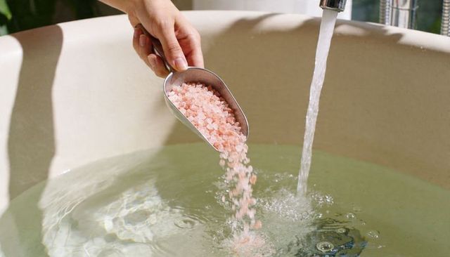 Hand pouring pink bath salts into freestanding tub with running faucet for spa