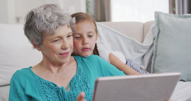Elderly woman and young girl using tablet while sitting on sofa in cozy living room. Ideal for promoting family bonding, multi-generational tech usage, and leisure activities. Useful for advertisements, blog posts, websites focusing on family relationships, technology integration in daily life, or elder and child interactions.