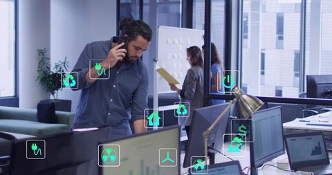 Talking male professional leaning at desk checking monitor with smartphone in open office