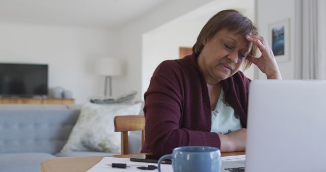 Senior woman sitting at a table with a laptop, looking stressed and tired, likely working from home. Suitable for use in topics related to remote work, senior employment, stress, technology use among elderly, and mental health.