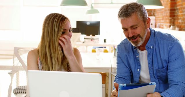 A professional man and woman sit together in a brightly lit modern office space, engaging in a friendly conversation while using a laptop and a tablet. Ideal for use in content highlighting teamwork, office productivity, modern work environments, or discussions about corporate culture and technology integration.