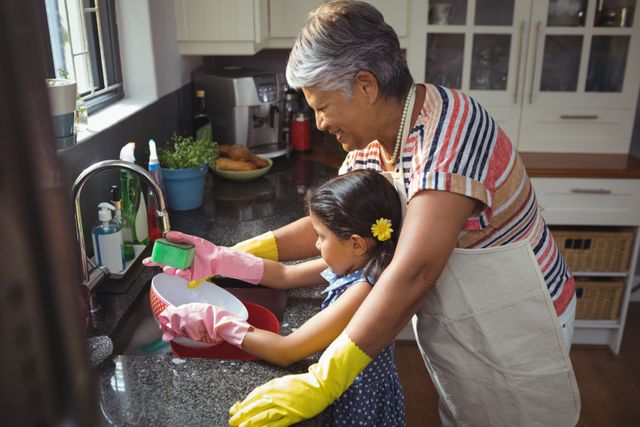 Grandmother and granddaughter washing dishes together in a modern kitchen. Both are wearing rubber gloves and smiling while cleaning a bowl. This image can be used to depict family bonding, intergenerational relationships, and domestic chores. Ideal for use in advertisements, family-oriented content, and articles about home life and family values.