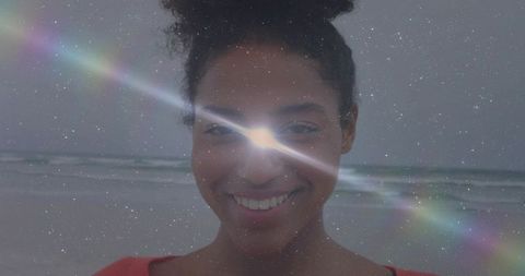 Smiling woman standing at ocean shoreline facing camera with rainbow lens flare, starfield