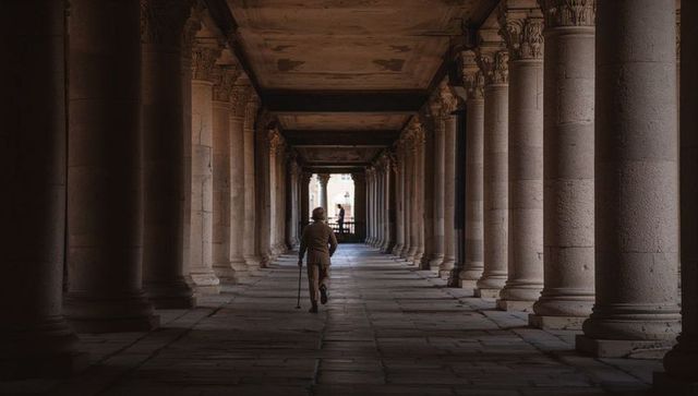 Walking elderly man through historic stone colonnade using cane, moody architectural corridor scene