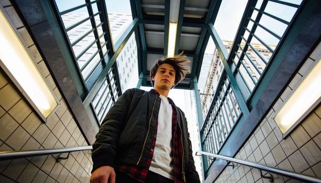 Windswept teen standing on urban staircase under glass canopy wearing bomber jacket low-angle pose