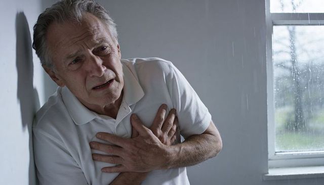 Senior man clutching chest in pain near rainy window portraying sudden heart attack distress