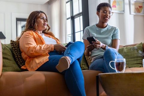 Diverse women relaxing on sofa holding smartphones, casual conversation at home