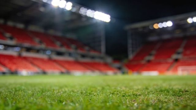 Foreground turf glistening under stadium floodlights at night with blurred red stands