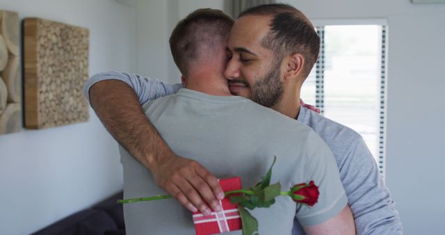 Two men hugging with affection, one holding a red rose and gift box behind his back, suggesting a romantic gesture or celebration. The image is ideal for use in campaigns or content focused on love, relationships, LGBTQ+ representation, Valentine's Day, or general themes of affection and happiness.