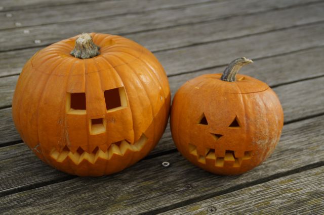Two carved pumpkins with jack-o'-lantern faces sitting on a wooden deck. Ideal for Halloween-themed articles, autumn decorations, social media posts sharing holiday spirit, and commercial use in advertisements promoting Halloween events or products.