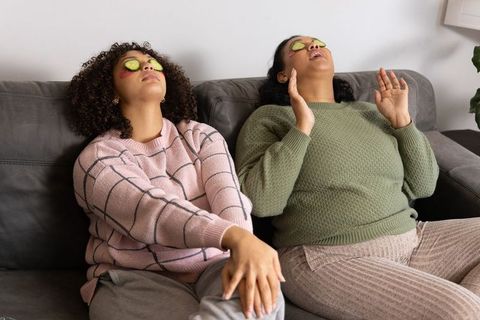 African american women relaxing on couch with cucumber slices and eye patches, cozy self-care