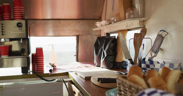 Shot captures inside of food truck focusing on counter with coffee machine, cups stacked, and basket of bread rolls. Ideal for themes related to street food, small business, mobile kitchen setups and food service environments.