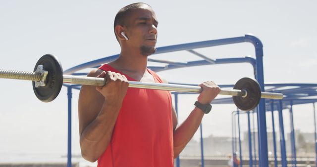 Focused biracial man lifting weight bar and wearing earphones at seaside. Sport, active lifestyle and technology.