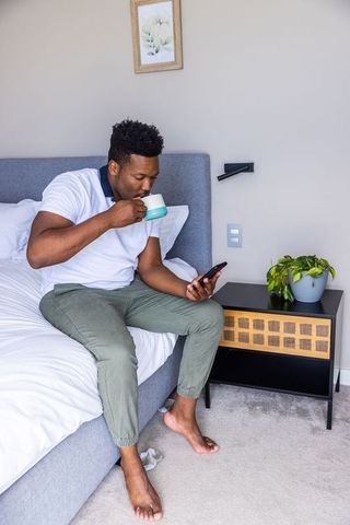 African american man sipping coffee and checking smartphone while sitting on modern bed