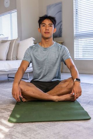 Young asian man practicing mindfulness on yoga mat in bright living room wearing smartwatch