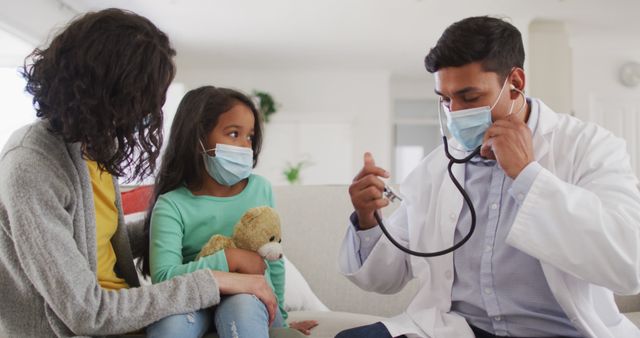 Hispanic male doctor holding stethoscope talking to mother and daughter at home wearing face masks. medical professional making patient home visit during coronavirus covid 19 pandemic.