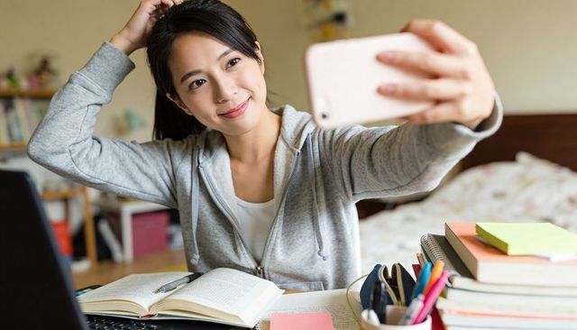 Young student taking selfie while studying at bedroom desk with laptop textbooks and phone