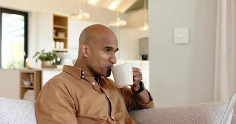 Man sipping coffee while working on laptop in sunlit living room with minimalist kitchen
