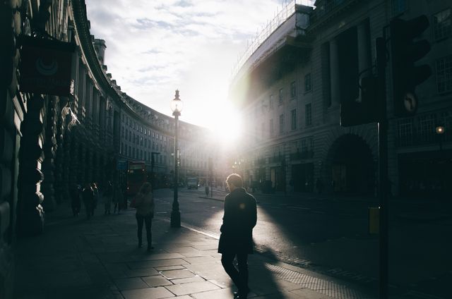 This image displays an early morning urban street scene with silhouetted pedestrians walking past elegant buildings. The rising sun creates a dramatic effect, illuminating the architecture and casting long shadows. This stock photo is ideal for use in travel brochures, city lifestyle articles, content focusing on morning routines, or urban planning presentations.