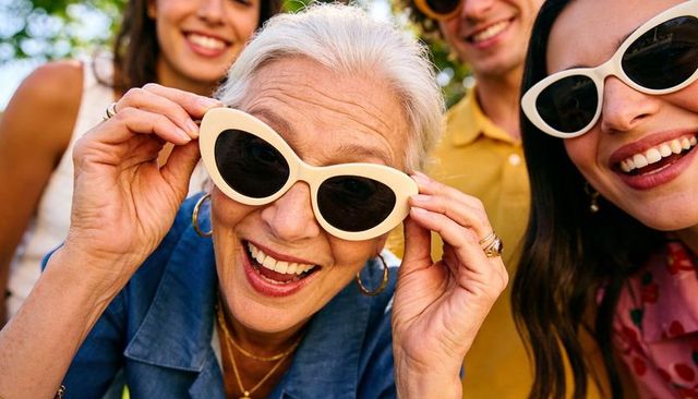 Joyful senior woman adjusting white cat-eye sunglasses with smiling friends in sunny park close-up