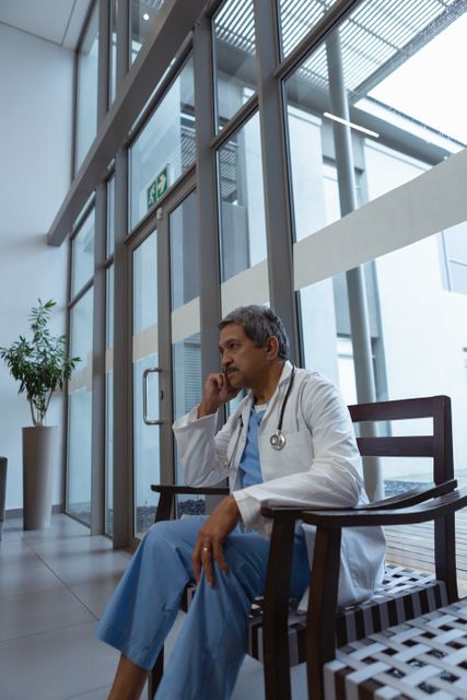 Side view of thoughtful mature male doctor with hand on face sitting on chair in lobby at hospital