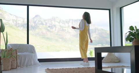 Woman in yellow jumpsuit standing at large window gazing at mountain view