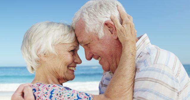 Elderly couple smiling and embracing on a beach on a sunny day. This image can be used for projects related to senior living, retirement communities, lifestyle, love, relationships, and travel advertisements. Ideal for brochures, websites, and social media content about active and happy senior life.