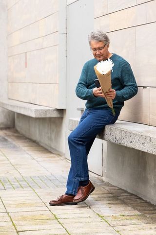 Senior man sitting on urban bench holding bouquet and checking smartphone, thoughtful moment