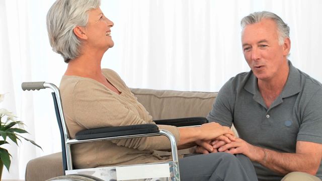 Senior couple engaging in conversation in a modern living room with natural light. The woman is sitting in a wheelchair while the man holds her hands, both smiling warmly. Suitable for use in healthcare, retirement and lifestyle contexts, emphasizing support, care and the beauty of elderly companionship.