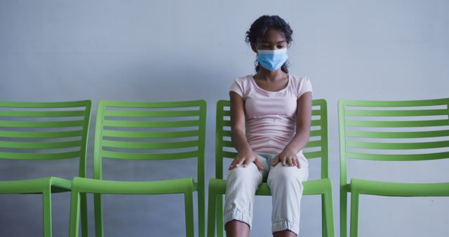 A young girl is sitting alone on a row of green chairs in a waiting area while wearing a face mask. Ideal for depicting themes related to healthcare, safety precautions, social distancing during the pandemic, and waiting in anticipation. Useful for illustrating concepts of childhood during COVID-19, patient anticipation, or serene healthcare environments.
